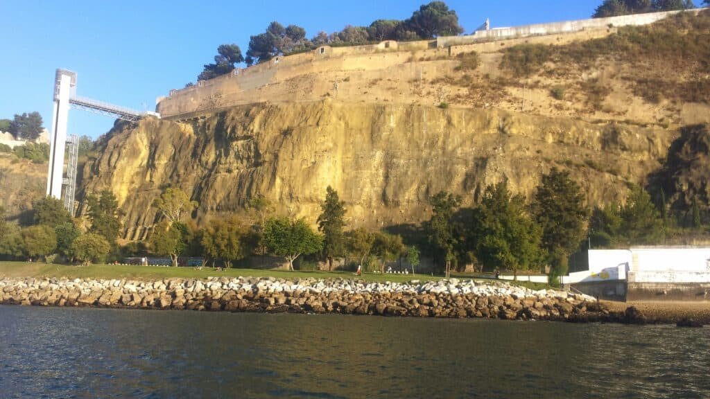 Jardim com uma falésia ao fundo e elevador panorâmico visto desde o rio Tejo